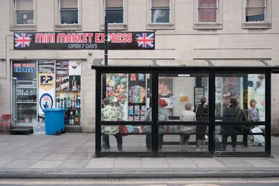 A bustling city bus stop in front of a mini market with people waiting and street advertisements.
