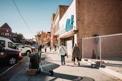 Two adults walking on a bright urban sidewalk lined with parked vehicles and shops.